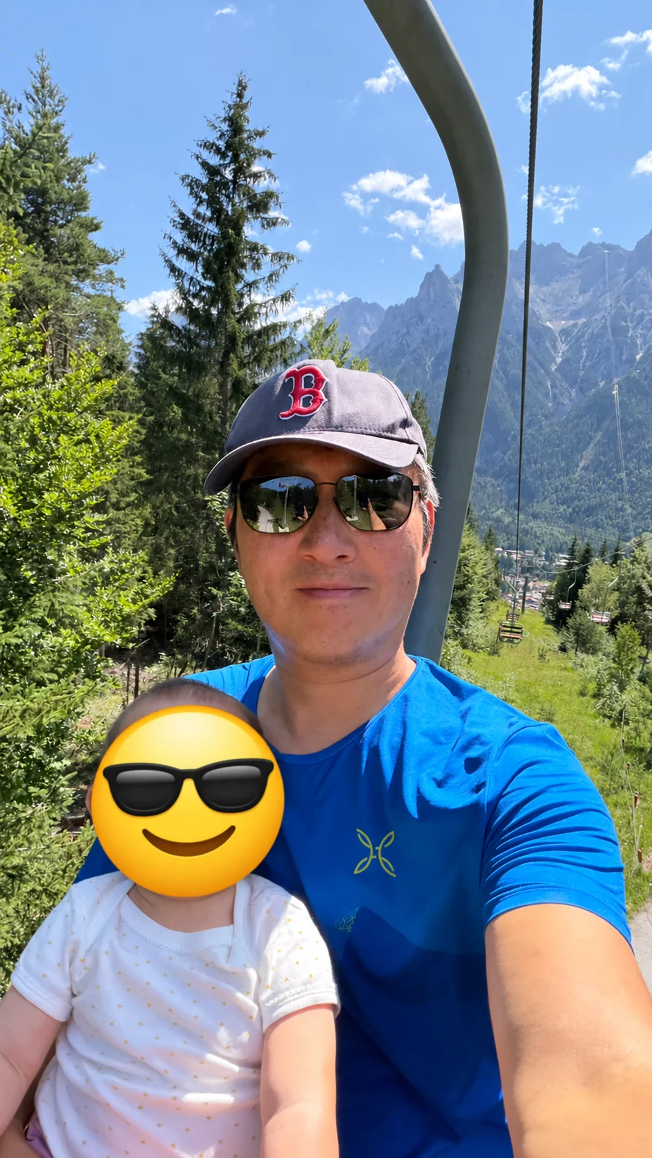 Ping mit seinem Kind im Sessellift vor einer Bergkulisse. Das Gesicht des Kindes ist mit einem Sonnenbrillen-Smiley verdeckt.
